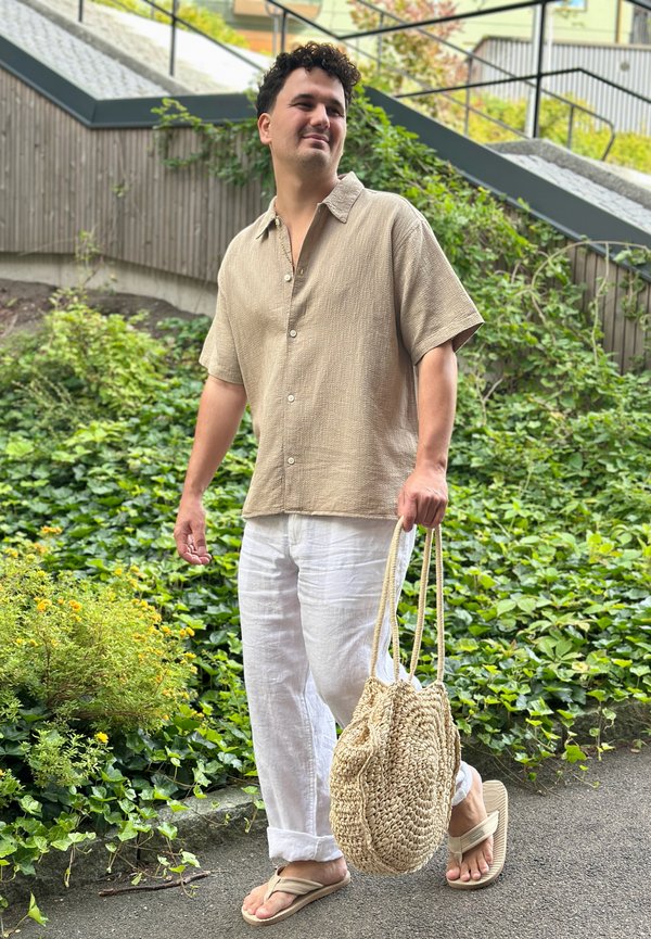 Homme portant une chemise beige, un pantalon blanc et des tongs, portant un sac fourre-tout tissé, se tenant à l'extérieur près de plantes vertes et d'escaliers.