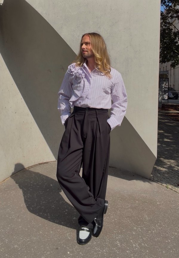 Personne aux longs cheveux blonds se tient contre un mur, portant une chemise blanche à volants, un pantalon large noir et des chaussures noires et blanches.