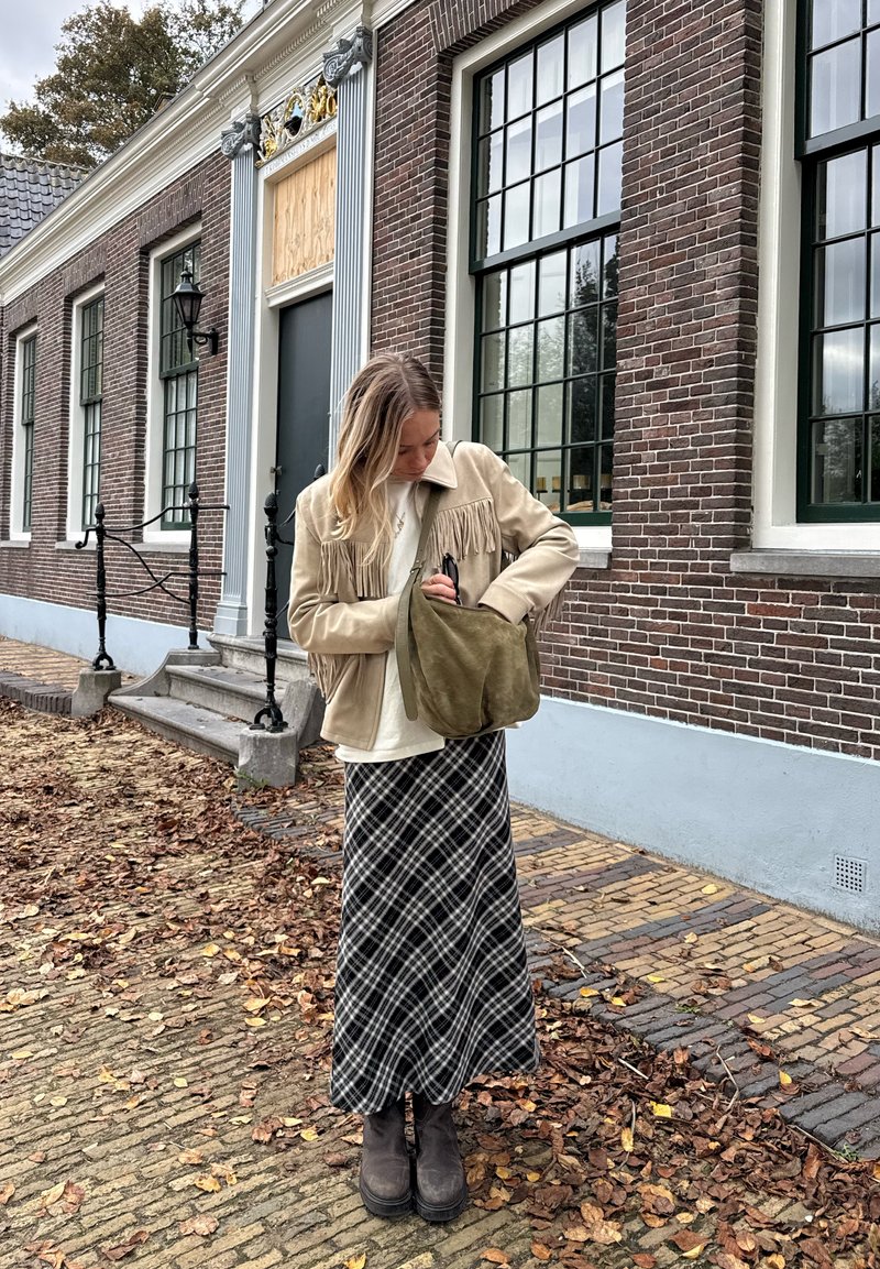 Vrouw in een beige franjesjack en een geruit rokje staat naast een bakstenen gebouw en kijkt in een groene suède schoudertas op een met bladeren bedekte stoep.