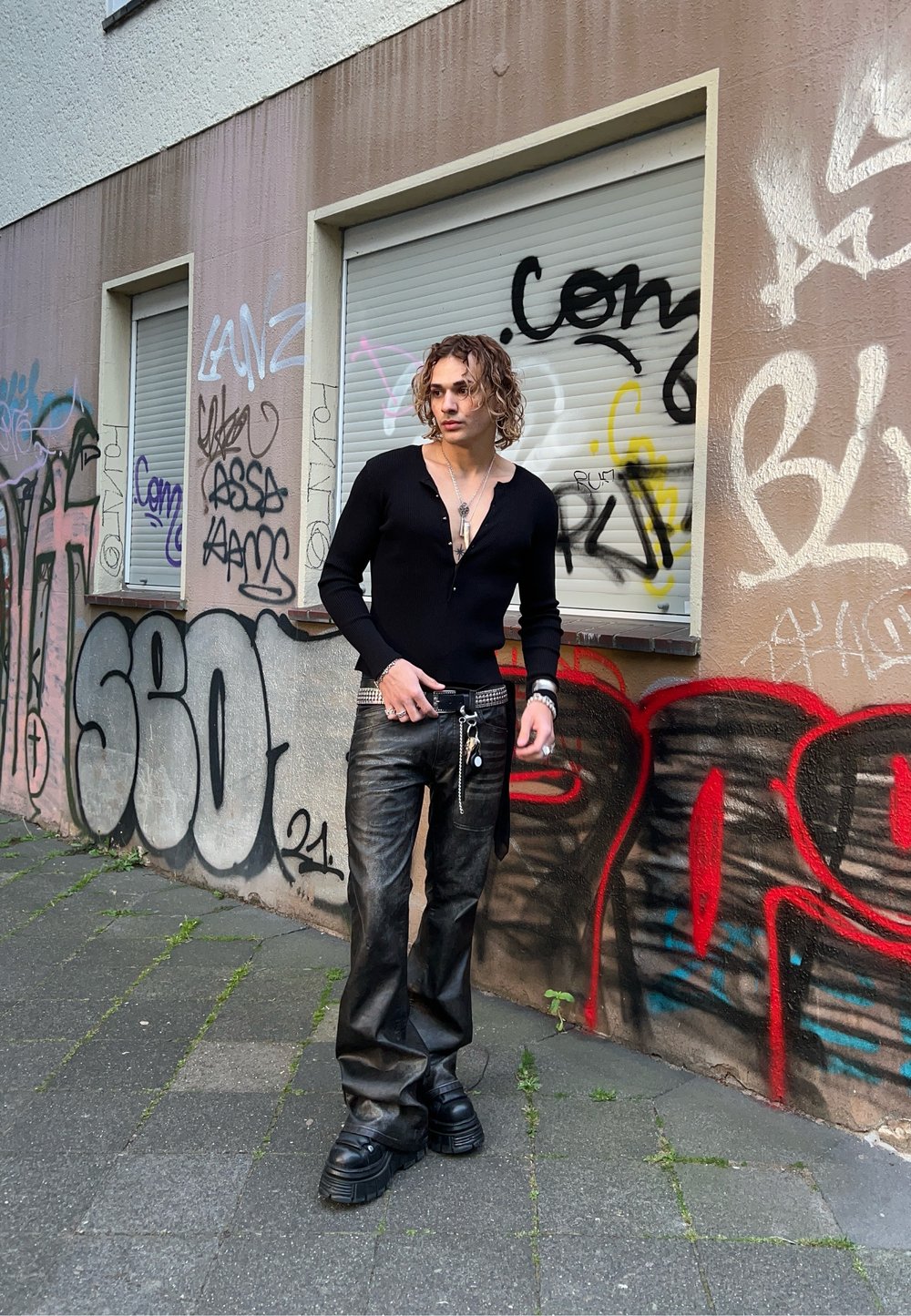 Μαύρος πουλόβερ με κουμπιά και βαθύ V-λαιμό, και φαρδιά μαύρα δερμάτινα παντελόνια. Χοντρά μαύρα παπούτσια και στρωματοποιημένα ασημένια κοσμήματα συμπληρώνουν την εμφάνιση.