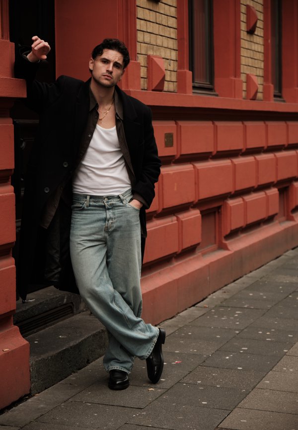 Muškarac u crnom kaputu, bijelom potkošulji i širokim plavim trapericama naslanja se jednom rukom u džep na trotoaru uz zgradu od crvene cigle u gradu.