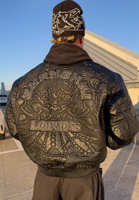 Jachetă bomber din piele neagră cu modele detaliate în relief. Prezintă textele „SHANGHAI” și „LORDS” cu accente metalice cu ținte pe spate.