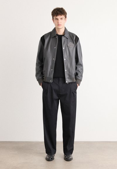 Chaqueta varsity de cuero negro con acentos grises, botones a presión y un parche con el logo. Lleva puesta sobre un suéter negro y combinada con pantalones negros holgados.