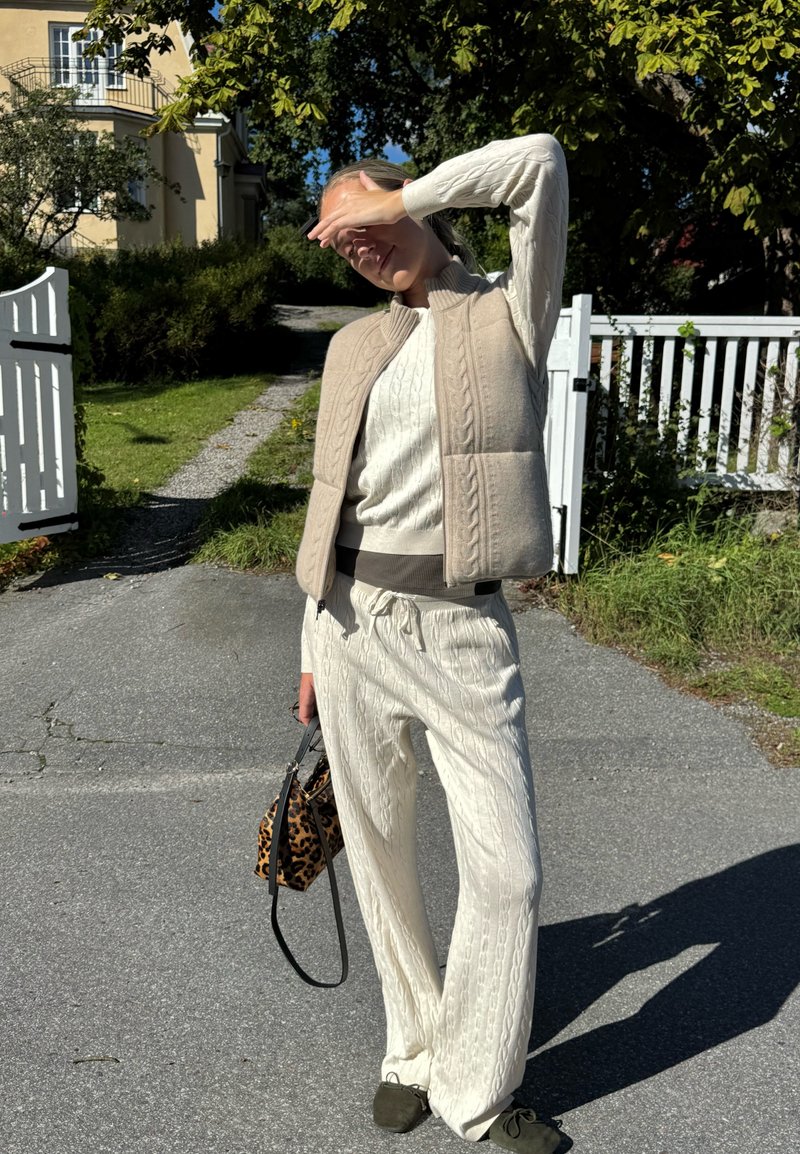 Gilet in maglia beige sopra un maglione crema con lavorazione a trecce, abbinato a pantaloni bianchi in maglia a trecce e scarpe verde oliva, mentre tiene una borsa con stampa leopardata.