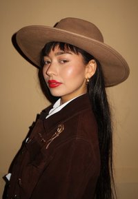 Donna con lunghi capelli neri che indossa un cappello marrone a tesa larga, una giacca marrone scuro con spille d'oro, rossetto rosso intenso e orecchini a cerchio dorati.