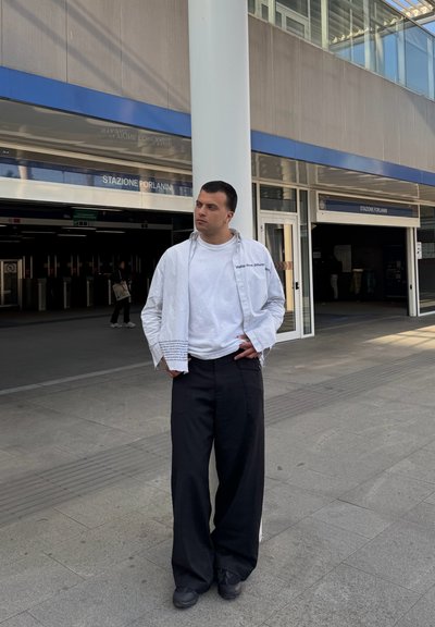 Persona vestida con una camiseta blanca debajo de una chaqueta ligera a rayas, pantalones anchos negros y zapatos negros, de pie frente a una estación de tren.