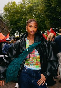 Zwarte oversized leren jas, kleurrijke gepatternde top, donkere spijkerbroek, teal gestructureerde tas en oorbellen met hoepels, geplaatst tegen een levendig buitenlandschap.