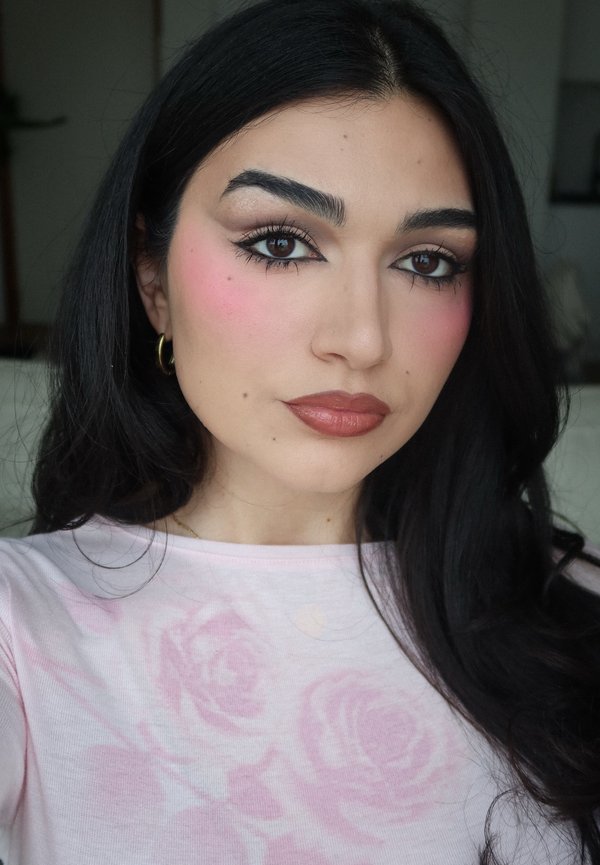 Vrouw met lang zwart haar, draagster van gouden oorringen in hoefijzervorm en een roze top met bloemenprint, met zachte roze blush en bruine lippenstift, kijkt naar de camera.