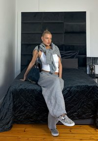 Jeune femme en pantalon de survêtement gris et débardeur blanc assise sur un lit noir, portant un sweat-shirt gris sur les épaules et des chaussures slip-on à carreaux.