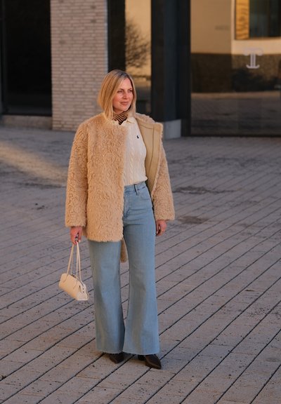 Mujer de pie al aire libre con un abrigo de piel sintética beige, suéter de punto blanco, vaqueros anchos azules, botas negras y sosteniendo un pequeño bolso crema.
