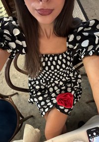 Donna che indossa un vestito a pois bianco e nero con maniche a sbuffo e una rosa di tessuto rosso mentre è seduta in un caffè.