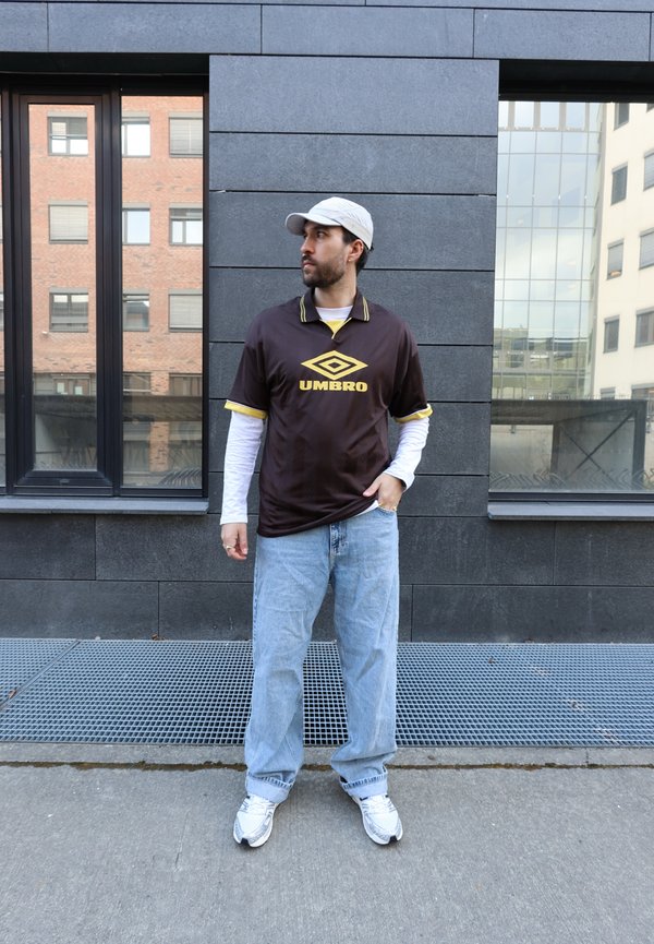 Jonge man met een lichte pet, een bruin Umbro-shirt over witte lange mouwen, losse lichtblauwe spijkerbroek en witte sneakers staat op een stedelijk trottoir.