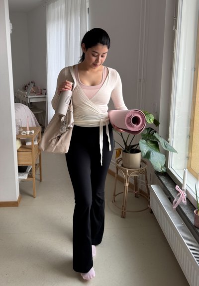 Estera de yoga rosa sostenida bajo el brazo; viste una blusa envolvente crema, camiseta sin mangas rosa claro y pantalones de campana negros; sostiene una botella de agua gris; ambiente interior.
