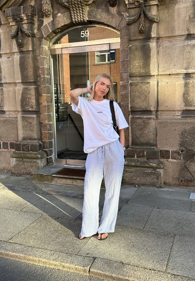 Camiseta blanca de gran tamaño con la palabra "Inglaterra" impresa, combinada con pantalones de pierna ancha a rayas azul claro. De pie frente a un edificio de piedra.