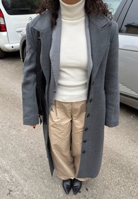 Personne portant un long manteau gris, un col roulé blanc, un pantalon beige, des chaussures noires à bout pointu, et portant un sac noir à franges, debout sur le trottoir près de véhicules.