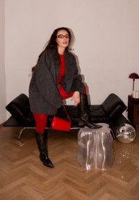 Femme en tenue rouge et bottes noires pose avec un pied sur un tabouret argenté dans une pièce avec un canapé noir, une boule disco et une petite lampe de table.