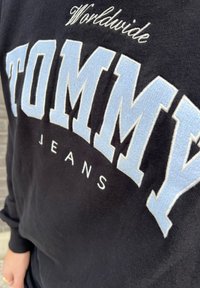 Fekete pulóver, amelyen feltűnően világoskék hímzett szöveg látható: "TOMMY" és alatta "JEANS", puha tapintású és bordás mandzsettákkal.