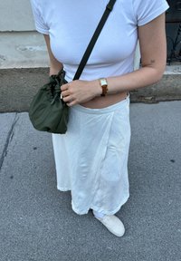Λευκή, ρέουσα φούστα και εφαρμοστό λευκό μπλουζάκι, συνδυασμένα με έναν ελκυστικό ελαιόχρωμο σάκο με κορδόνι και εκρού παπούτσια σε γκρι πεζοδρόμιο.