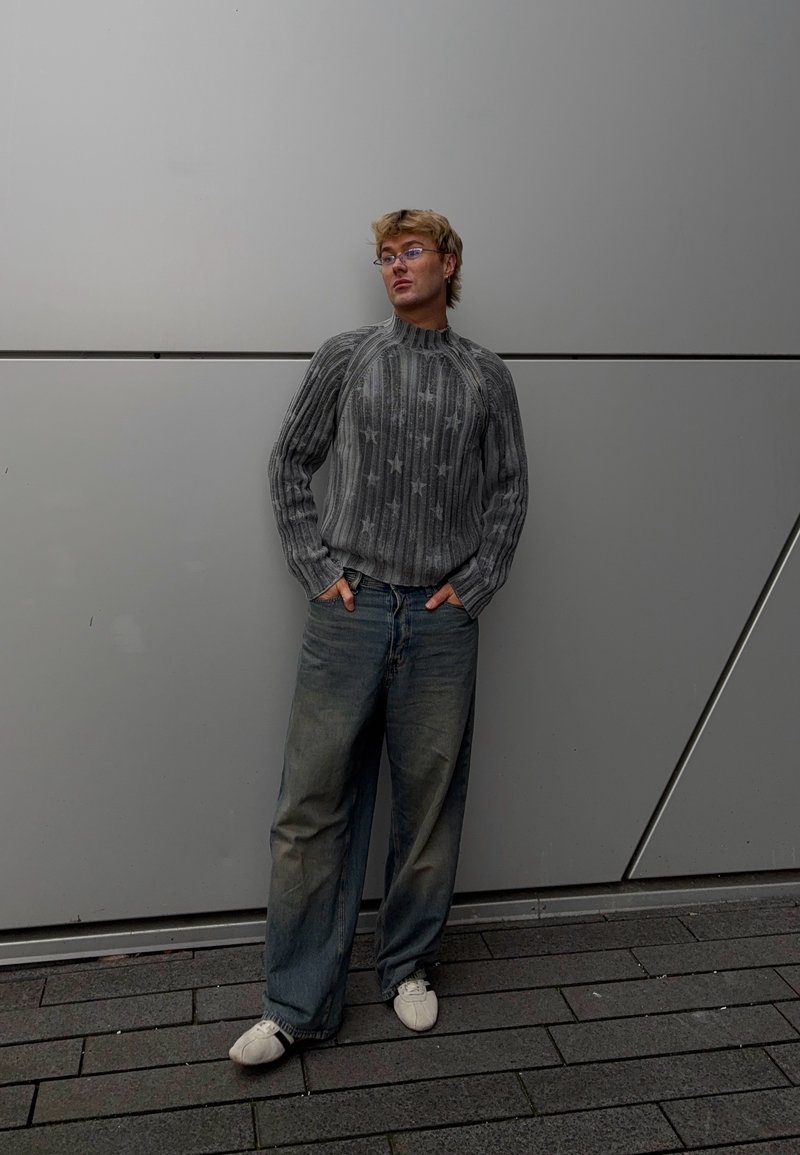 Jeune personne avec des lunettes appuyée contre un mur de panneaux gris, portant un pull gris à motifs d'étoiles, un jean ample et des chaussures blanches.