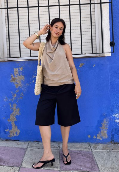 Top drapeado beige, pantalones cortos anchos negros, sandalias de tacón negras y un bolso de mimbre. El fondo es azul brillante con un marco de ventana blanco.