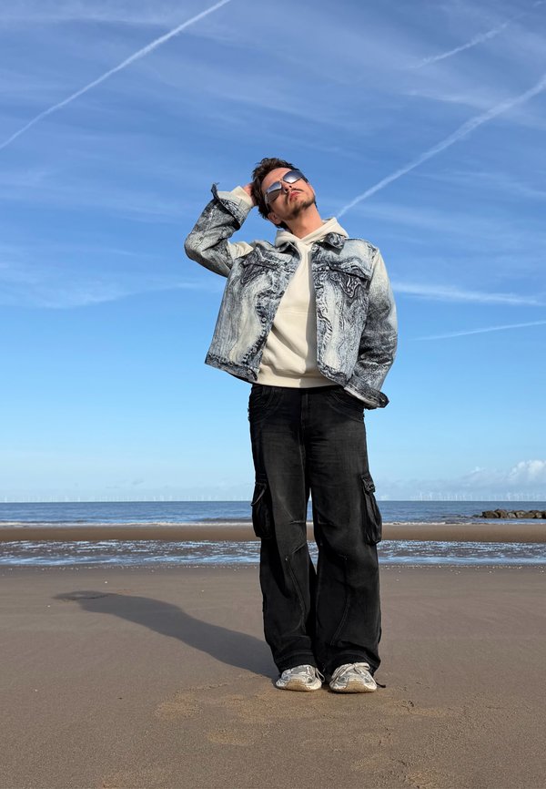 Joven con gafas de sol que lleva una chaqueta vaquera, una sudadera con capucha y pantalones cargo negros, está de pie en una playa de arena cerca del océano bajo un cielo despejado.
