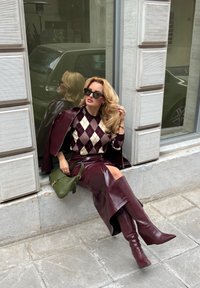 Donna con capelli biondi che indossa occhiali da sole, maglione a rombi, giacca di pelle bordeaux e gonna, e stivali alti, seduta sul marciapiede accanto a una finestra.