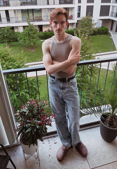 Top sin mangas de color gris claro, jeans de mezclilla azules, zuecos marrones. De pie en un balcón con plantas en macetas y un paisaje verde de fondo.