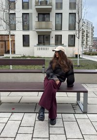 Γυναίκα με καπέλο, σκούφο πουλόβερ και μπορντό παντελόνι κάθεται σε παγκάκι σε αστικό πάρκο με μοντέρνα διαμερίσματα στο φόντο.