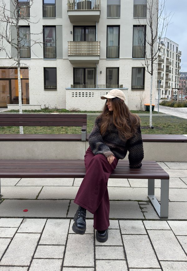 Vrouw met een pet, een donkere trui en bordeauxrode broek zittend op een bankje in een stadspark, met moderne appartementencomplexen op de achtergrond.