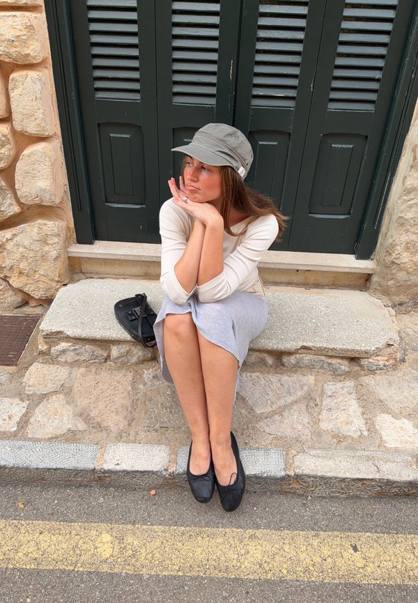 Une femme portant une casquette grise est assise sur des marches en pierre devant des volets vert foncé, le menton posé sur ses mains. Un sac à main noir est posé à côté d’elle.