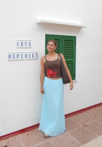 Ung kvinne i brun tanketopp og lang lyseblå skjørt står ved siden av en hvit vegg med grønn skodde vindu og «Casa Benedikt» skilt.