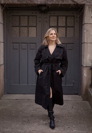 Abrigo de trench negro con cintura ceñida, gran cuello y botones, combinado con botas negras hasta la rodilla, contra un fondo de puerta gris.