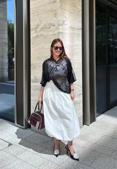 Top de malla negro con encaje y logo de Adidas, falda blanca fluida, zapatos de tacón negros con acentos de flores y un bolso burdeos con rayas blancas.