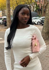 Vaaleanpunainen punottu käsilaukku, jossa solmittu yläosa ja kultaiset yksityiskohdat. Pitkähihainen valkoinen mekko, jossa poimuttunut pinta. Ulkoilmassa kaupunkimaisemassa.