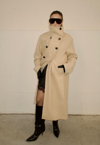 Persona con un abrigo beige cruzado de doble botonadura y cuello alto, falda de encaje negra, guantes negros, botas altas negras y grandes gafas de sol oscuras.