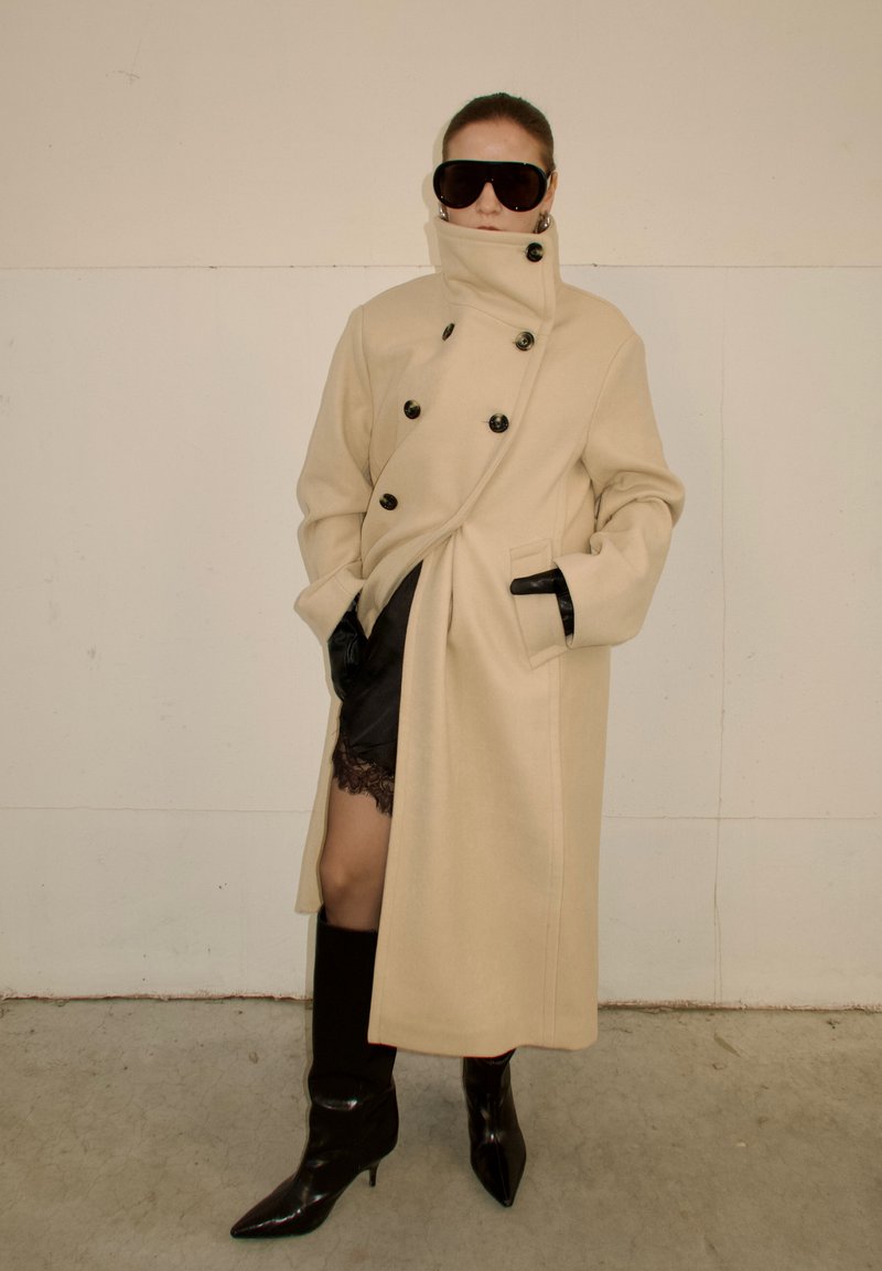 Personne portant un manteau beige à double boutonnage avec un col haut, une jupe en dentelle noire, des gants noirs, des bottes noires hautes et de grandes lunettes de soleil foncées.