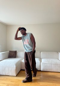 Mann mit dunkler Haut, der einen ärmellosen hellblauen Pullover und dunkle Hosen trägt, steht auf einem Holzboden vor einem weißen Ecksofa.