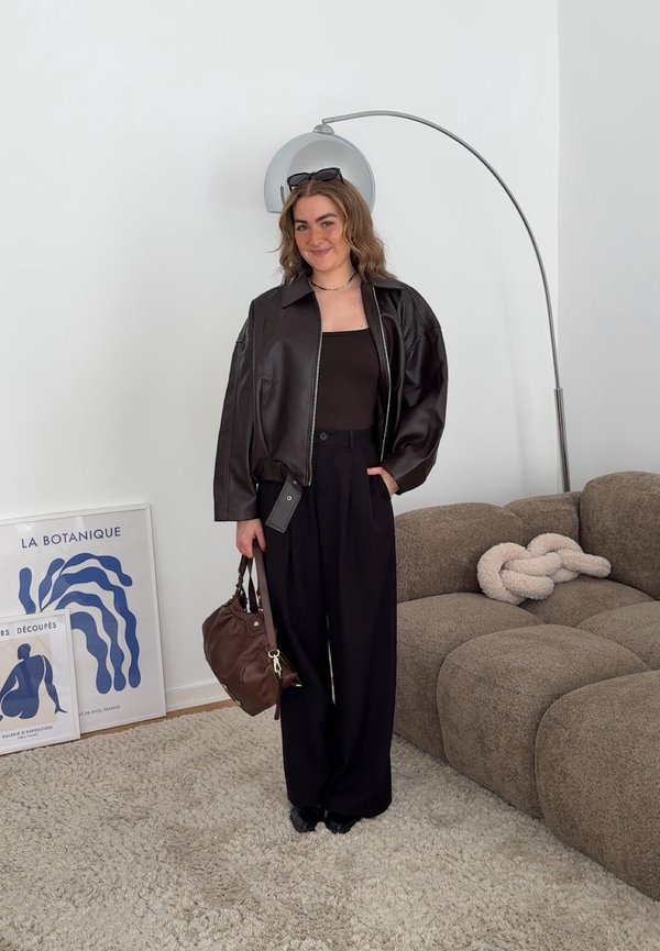 Junge Frau in schwarzer Lederjacke und Hose hält eine braune Tasche, steht lächelnd neben einem braunen Sofa und einem blau gerahmten Kunstwerk an einer weißen Wand.