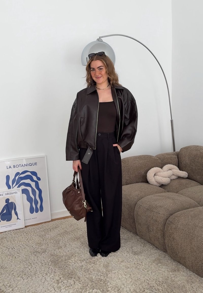 Junge Frau in schwarzer Lederjacke und Hose hält eine braune Tasche, steht lächelnd neben einem braunen Sofa und einem blau gerahmten Kunstwerk an einer weißen Wand.
