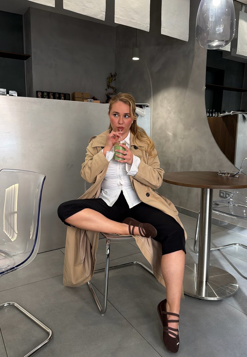 Frau in beige Mantel und schwarzen Hosen sitzt mit übergeschlagenen Beinen auf einem Stuhl und trinkt einen grünen Smoothie in einem modernen Café mit grauen Wänden und transparenten Stühlen.