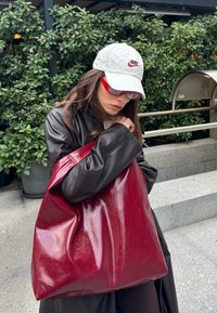 Femme portant une casquette blanche Nike et des lunettes de soleil rouges tient un grand sac fourre-tout rouge brillant, vêtue d'un manteau en cuir foncé à l'extérieur près de plantes.