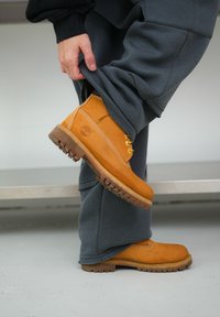 Κίτρινη δερμάτινη μπότες αστραγάλου με καουτσούκ σόλα, που διαθέτει χρυσές οπές, λευκή ραφή και υφή. Φοριέται με γκρίζες φόρμες.