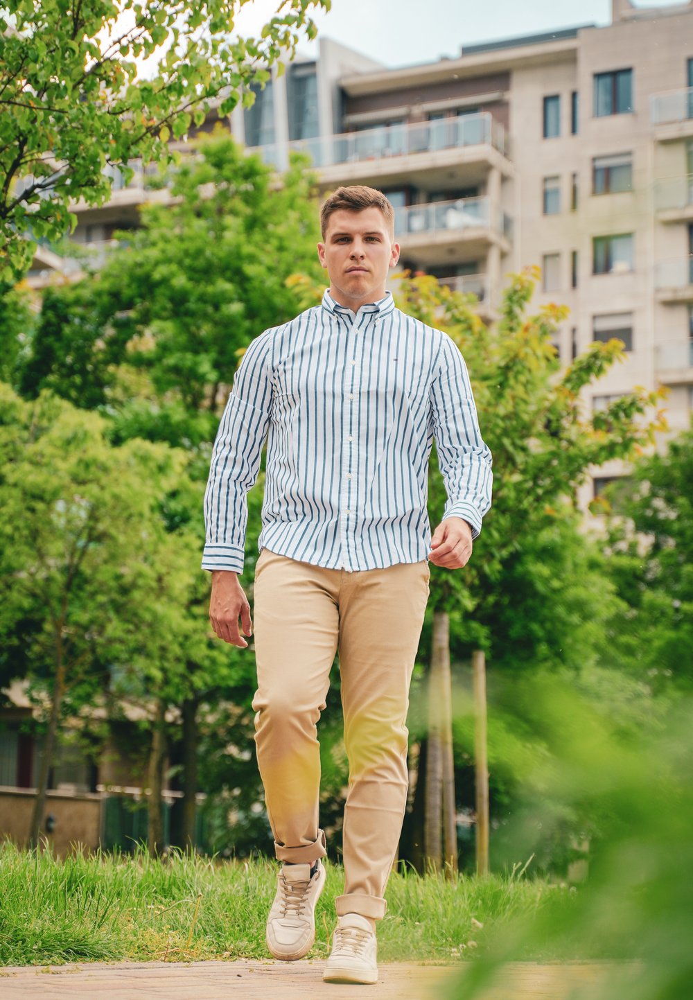 Vit skjorta med blå ränder, beige byxor och beige sneakers. Modell som går på en gångväg med grönska och moderna byggnader i bakgrunden.