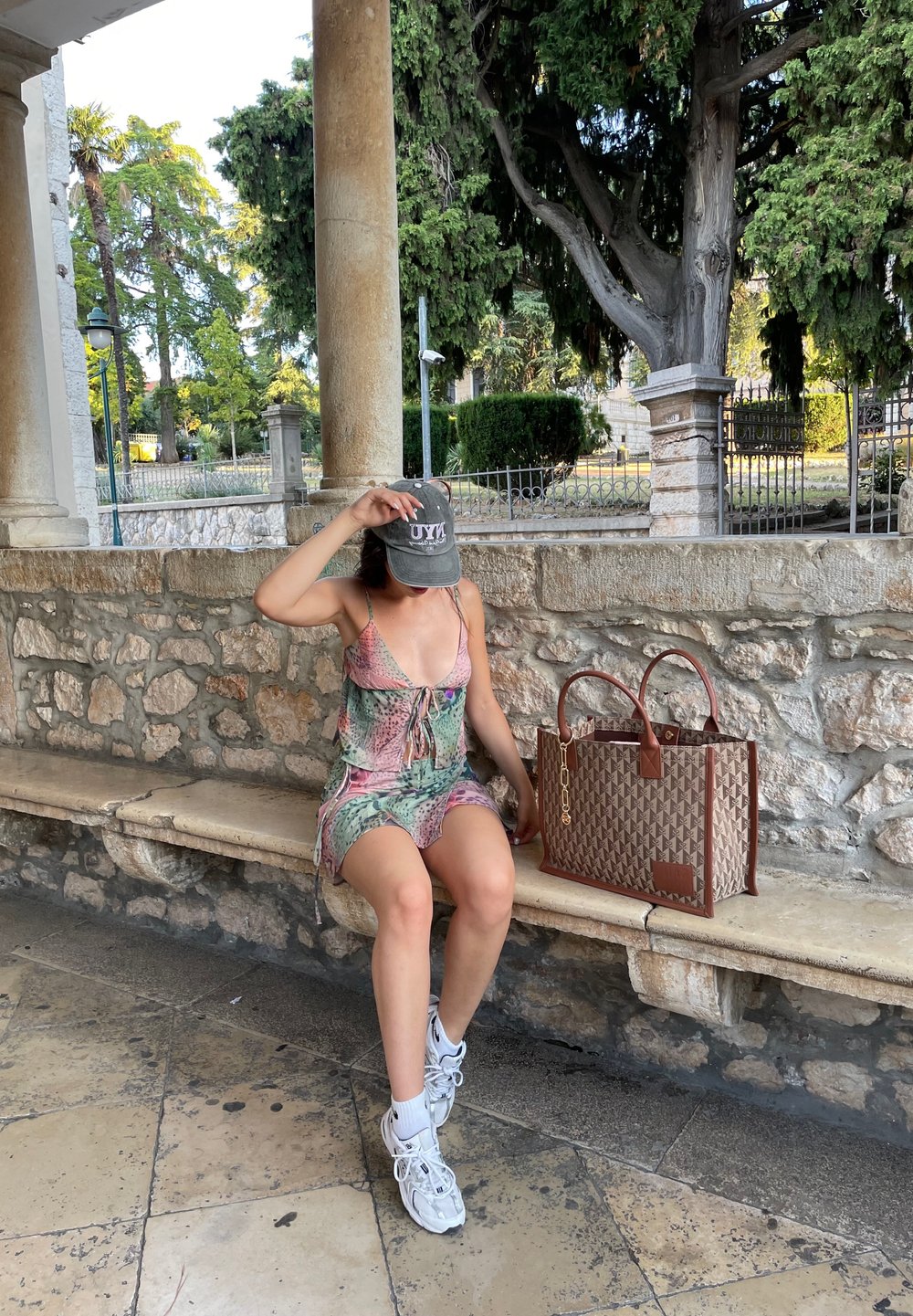 Blomstermønstret kjole med justerbare stropper, i nyanser av rosa og grønn. Brun geometrisk veske med kjededetaljer. Hvite sneakers og caps.