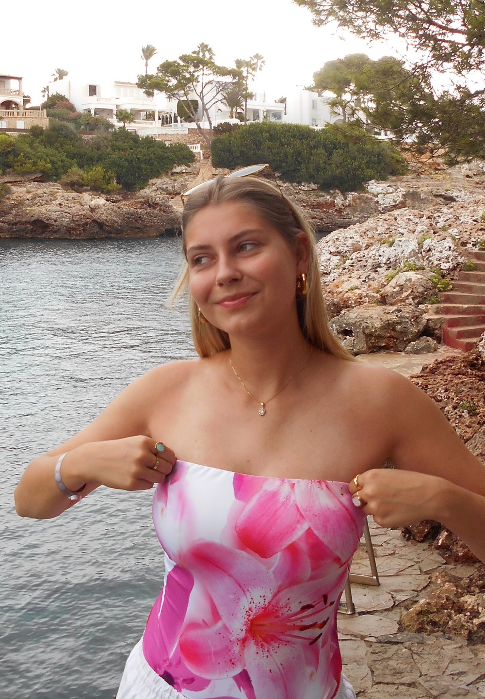Jonge vrouw met blond haar, gekleed in een strapless roze bloemen top, past deze aan bij de rotsachtige kustlijn met water en witte gebouwen op de achtergrond.