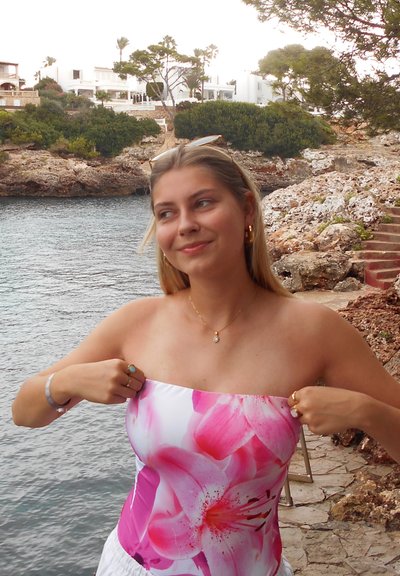 Joven mujer con cabello rubio que lleva un top floral rosa sin tirantes, lo ajusta junto a la costa rocosa con agua y edificios blancos de fondo.