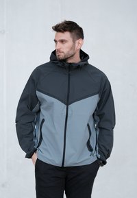 Kurtka Nike w kolorze czarnym i jasnoszarym, z zamkiem błyskawicznym z przodu, regulowanym kapturem oraz dwoma bocznymi kieszeniami, wykonana z gładkiego, teksturowanego materiału.