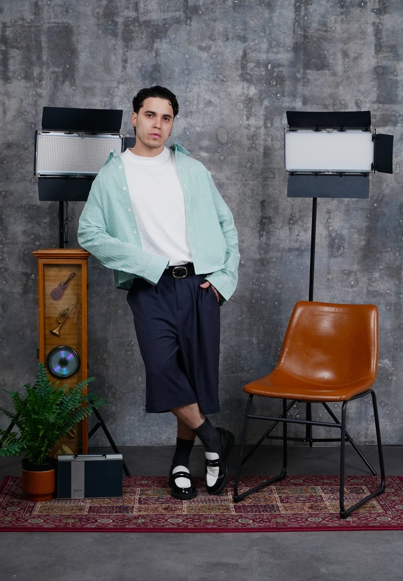 Jonge man in casual kleren leunt tegen een retro houten muziekmeubel, naast studiolampen, een leren stoel en een kamerplant op een patroonTapijt.