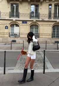 Mustad põlveni ulatuvad kummisaapad valge äärisega, kombineeritud kreemja kampsuni ja valgete lühikestega; käes lillekimp. Taustal kiviplaatidega kõnnitee.