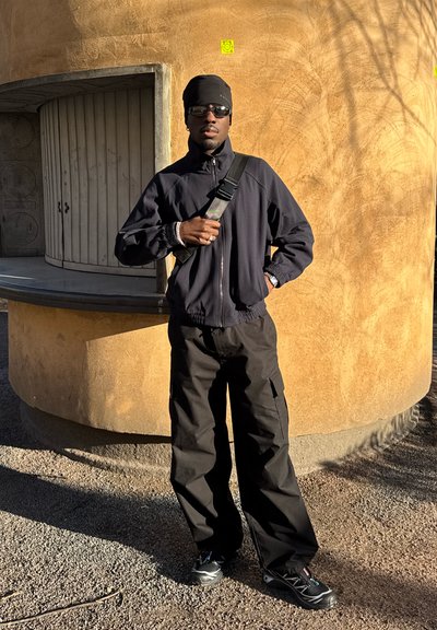 Hombre con chaqueta negra, pantalones, gorro y gafas de sol está de pie al aire libre frente a una pared curva texturizada con una ventana de madera cerrada.